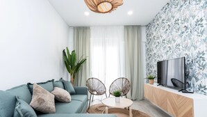 Een moderne woonkamer met een modulaire bank, een ronde tafel met twee stoelen, een televisie op een houten kast en een groot raam met transparante gordijnen.