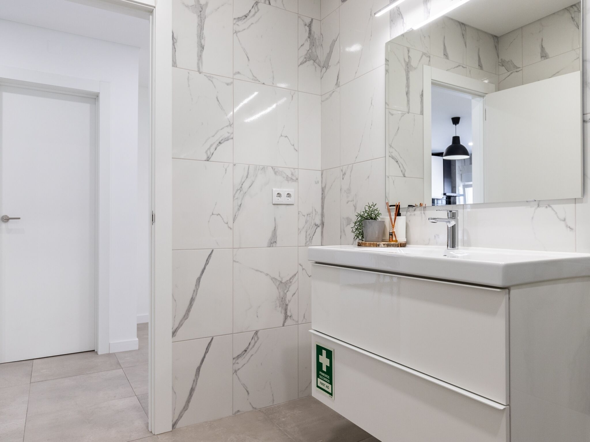 Ein modernes Badezimmer mit weißen Marmorfliesen, einem Waschbecken mit Spiegel und einem Erste-Hilfe-Schild an der Wand.