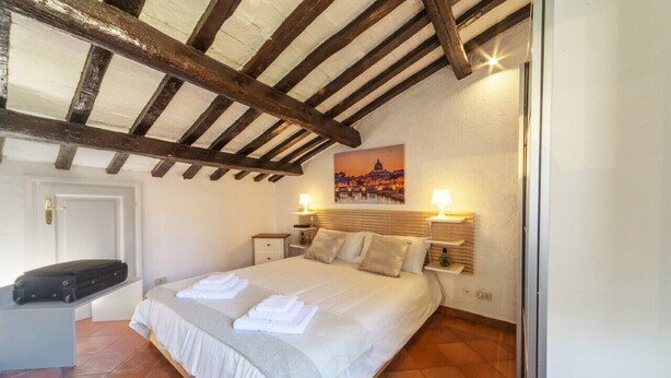 A bedroom with a bed, bedside lamps, a painting, and a suitcase on a table. - [Trastevere] Cozy & Charme Apartment in Rome (Roma)