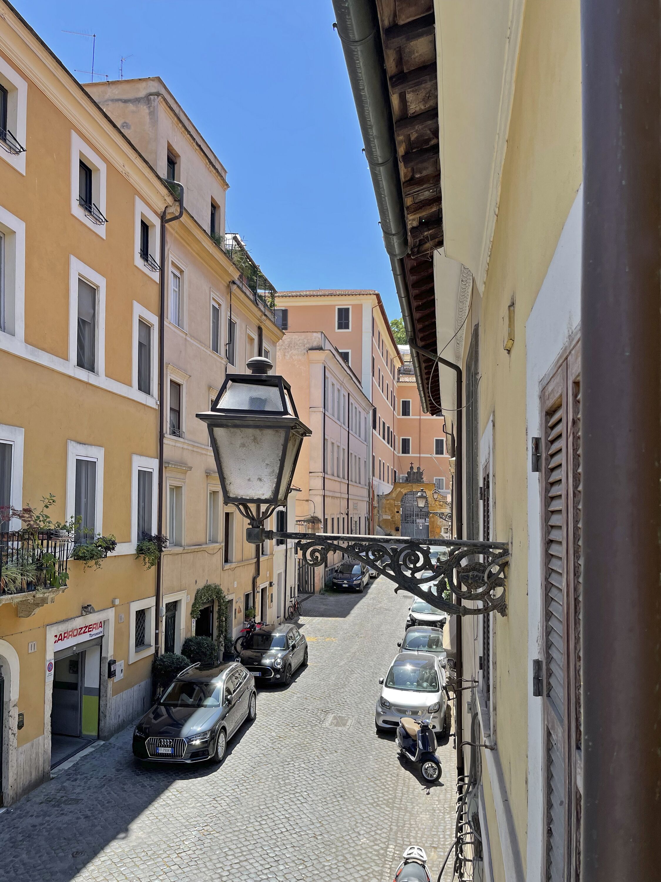 Una stretta strada acciottolata fiancheggiata da edifici a più piani, auto parcheggiate e un lampione.