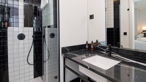 Ein modernes Badezimmer mit einer schwarz-weißen Dusche, einem weißen Waschbecken mit schwarzer Arbeitsplatte und einem großen Spiegel, der ein Schlafzimmer reflektiert.