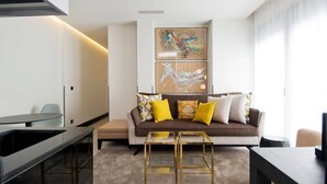 Ein modernes Wohnzimmer mit einem braunen Sofa, gelben Kissen und einem Glastisch. An der Wand hängt ein Gemälde, und es gibt einen Esstisch mit Stühlen.