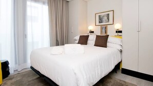 Ein ordentlich gemachtes Bett mit weißen Leinen und zwei Kissen, ein Nachttisch mit Lampe und ein großes Fenster mit durchsichtigen Vorhängen.