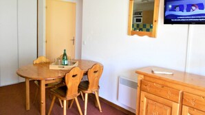 A room with a wooden dining table, two chairs, a small cabinet, and a television mounted on the wall.
