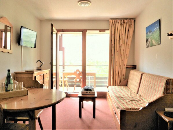 A room with a wooden table, a sofa, a television, and a window with curtains.