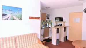 A compact kitchen with a microwave, coffee maker, and a small refrigerator. There is a wooden table and a sofa in the living area.