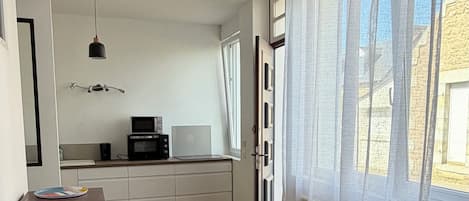 Ein geräumiges Zimmer mit Holzboden, einer Küchenzeile mit Mikrowelle, einem Esstisch und einem großen Fenster mit Vorhängen.
