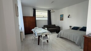 Quarto pequeno e bem iluminado com cama, sofá, mesa de jantar com cadeiras, guarda-roupa de madeira e janela com cortinas.