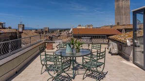 Terrassa a la teulada amb una taula rodona i seients, una planta en test i una vista de l'horitzó urbà amb un edifici alt al fons.