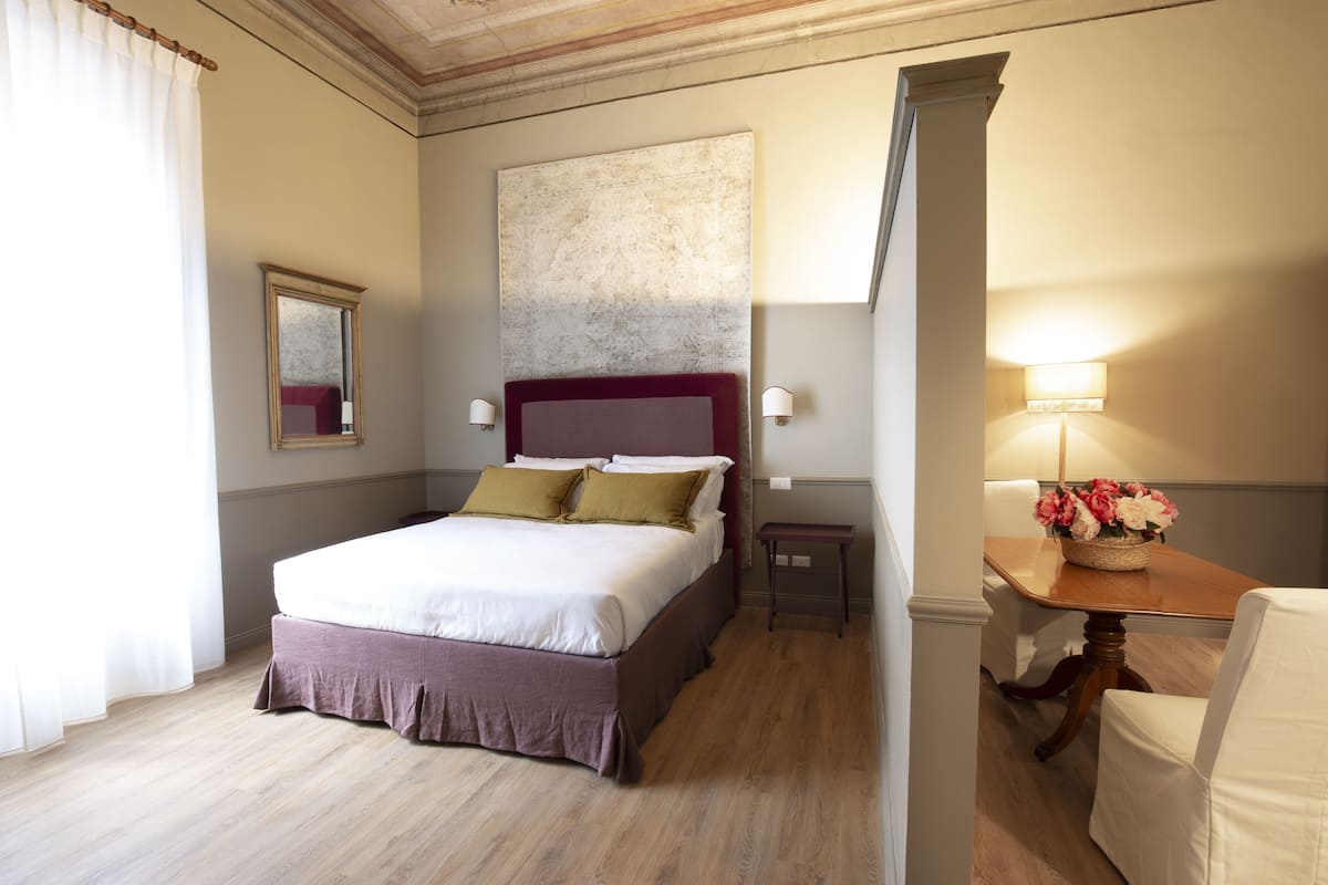 A hotel room with a large bed, a bedside table with a lamp, a small table with flowers, and a mirror on the wall.