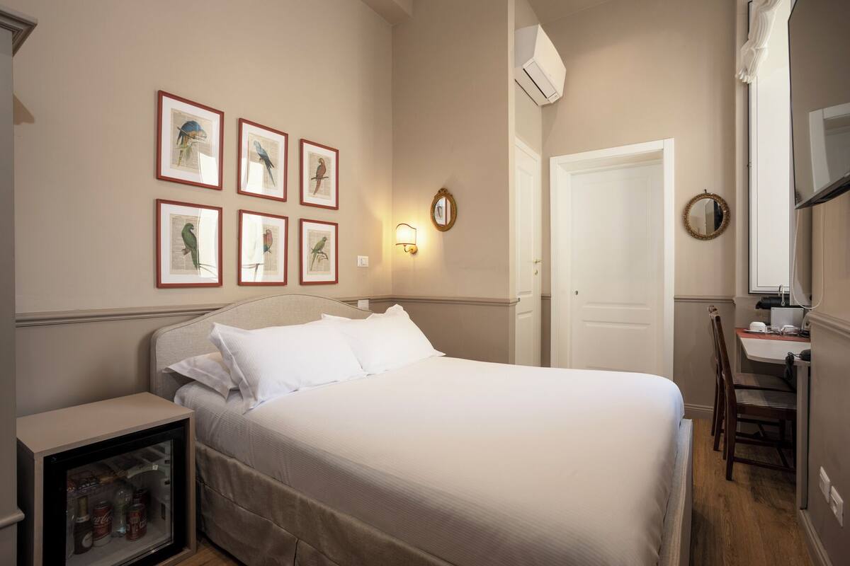 A hotel room with a bed, a desk, a chair, a refrigerator, and wall-mounted artwork.