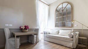 A living room with a sofa, a table, and a chair. There is a window with curtains, a mirror on the wall, and a lamp on a side table. - Hotel Donna Francesca (Rome)