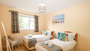 A hotel room with two beds, a window with curtains, a painting of beach chairs, a white chair, and a wooden floor. - Sunny Haven (Weymouth)