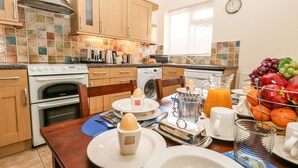 A modern kitchen with wooden cabinets, a built-in oven, a washing machine, and a dining area with breakfast items. - Sunny Haven (Weymouth)