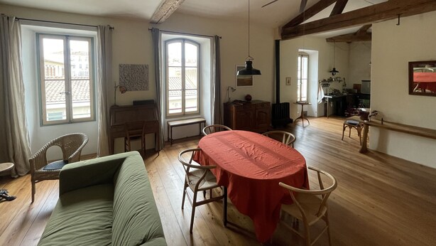 Ein geräumiges Wohnzimmer mit Esstisch, einer Couch und einem Deckenventilator. Große Fenster und ein Blick ins Freie sind vorhanden.