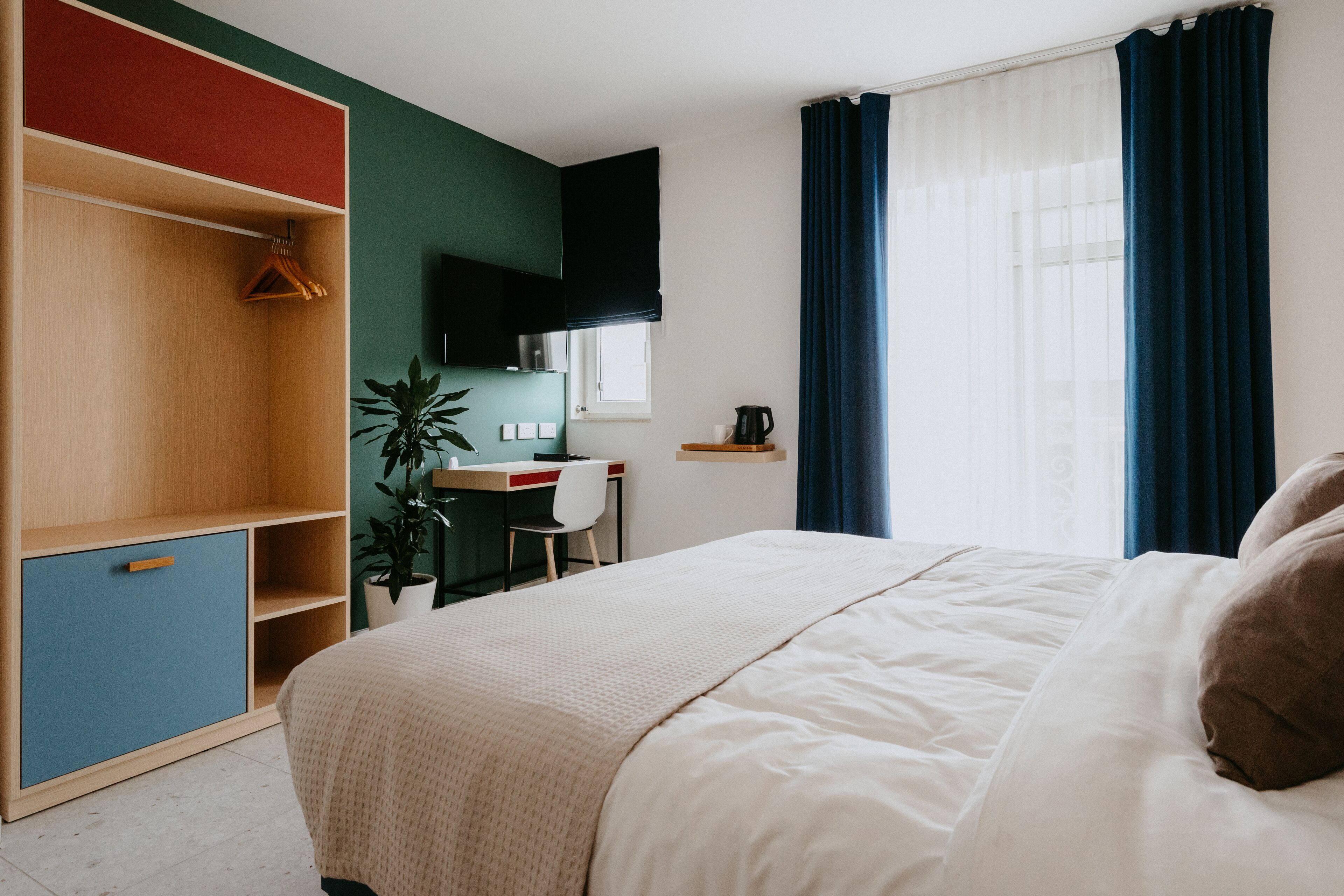 A bedroom with a bed, a desk, a chair, a plant, and a wardrobe.