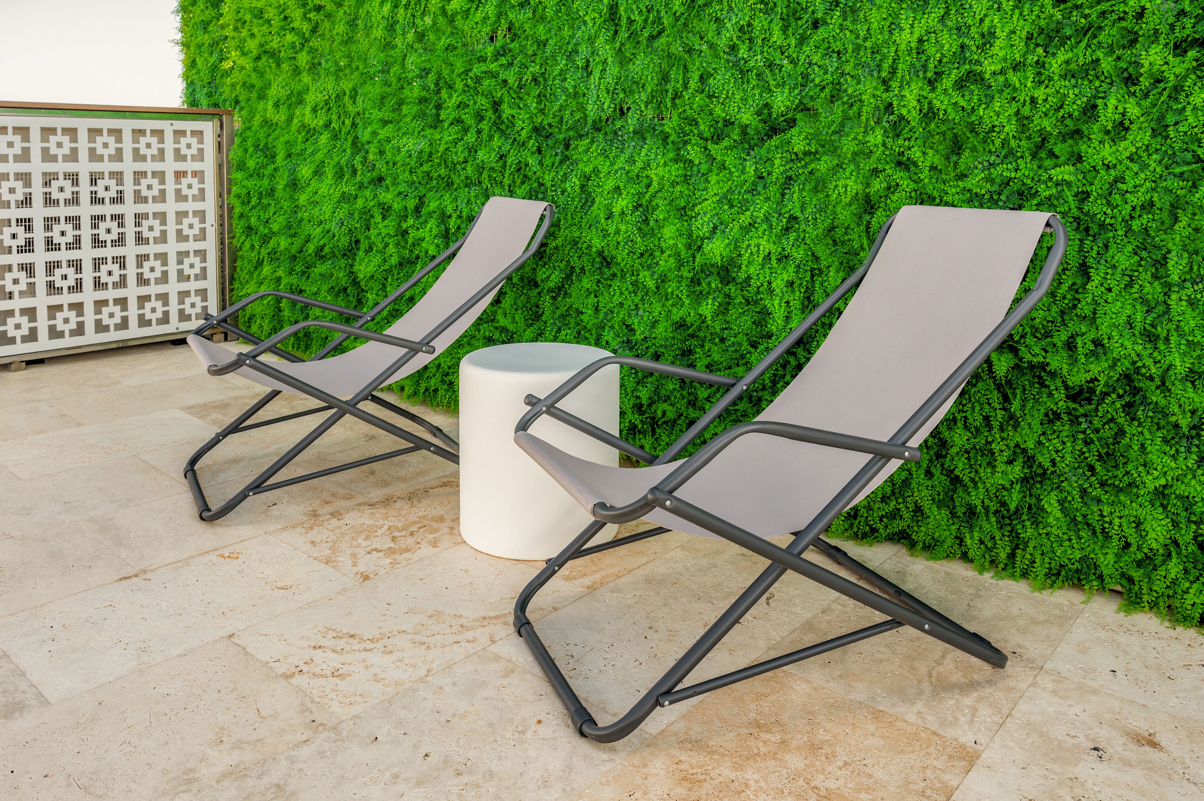 Two lounge chairs with metal frames and grey cushions, positioned on a tiled patio with a green hedge wall in the background.