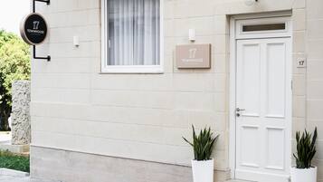 A townhouse with a white door, a window with white curtains, a signboard with the townhouse name, and two potted plants.