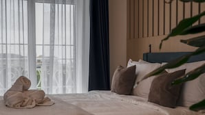 Een net opgemaakt bed met een beige deken en kussens, een raam met dunne gordijnen en uitzicht op groen.