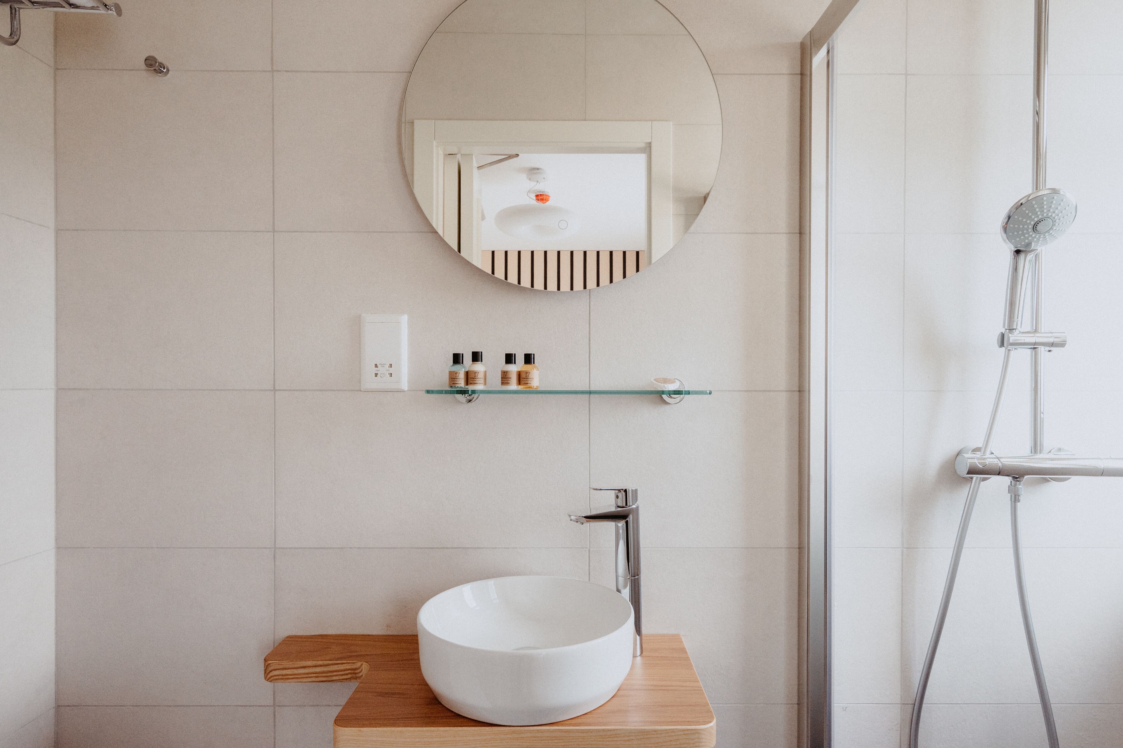 A modern bathroom with a round mirror, a wooden vanity, a white sink, and a glass shower enclosure.