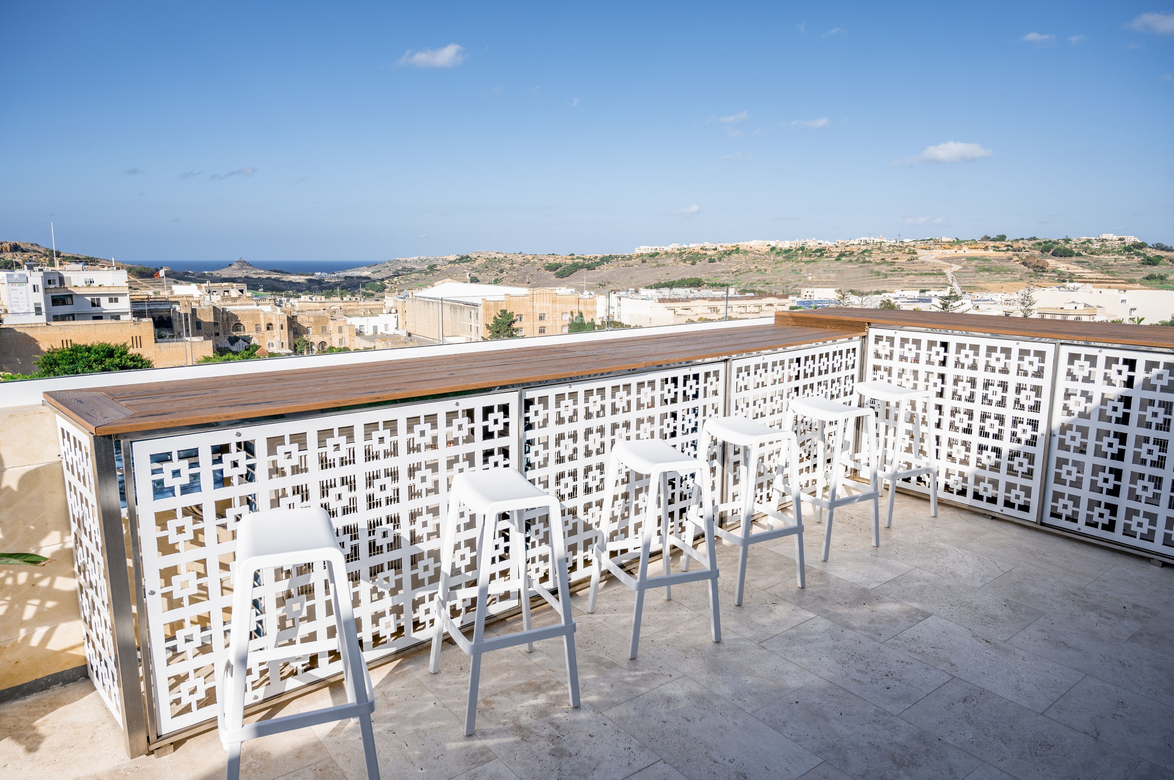 A rooftop terrace with a wooden bar and white bar stools, offering a view of the city and hills.