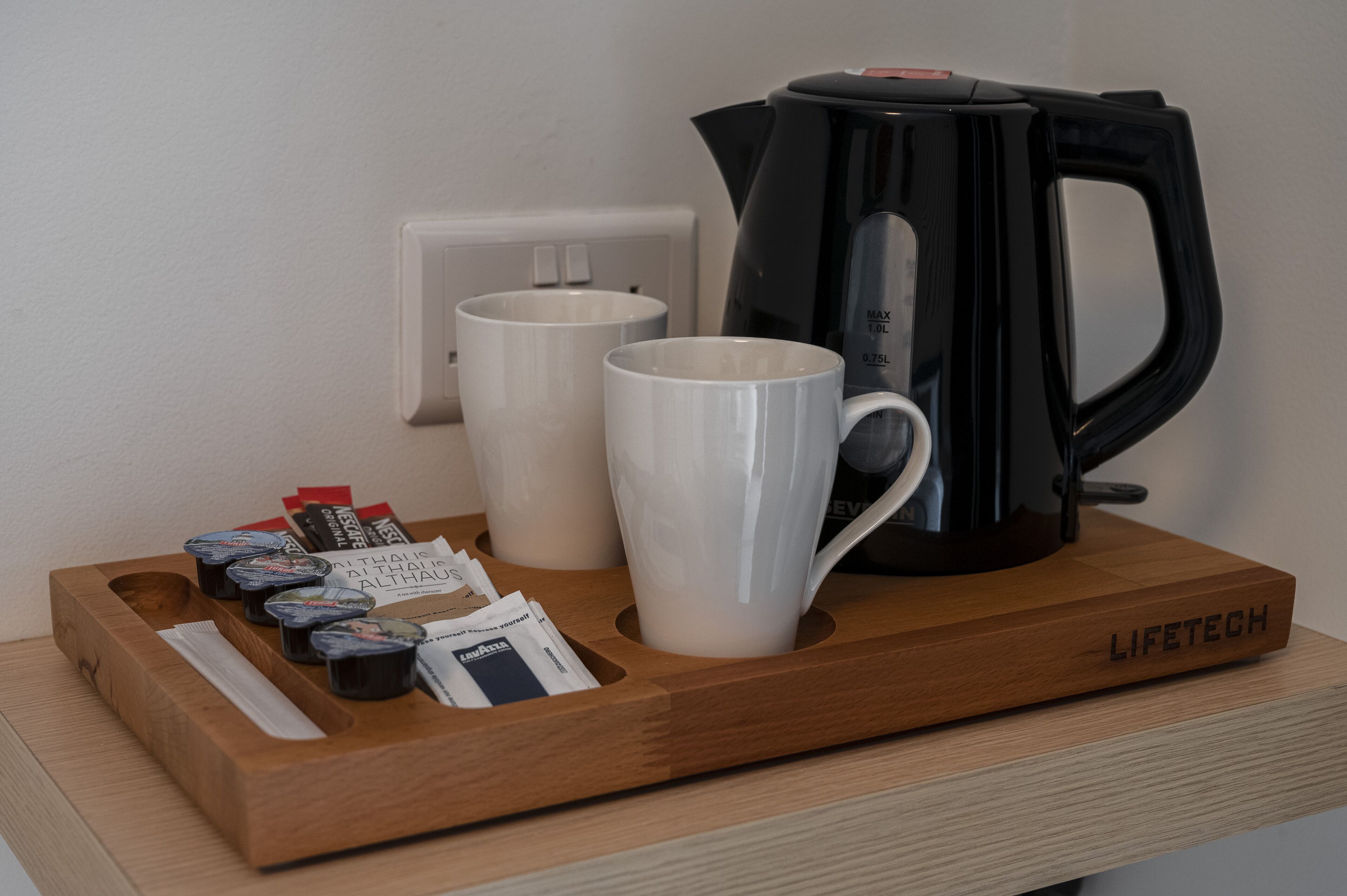 A coffee station with a black coffee maker, two white mugs, and a tray with coffee pods and a newspaper.
