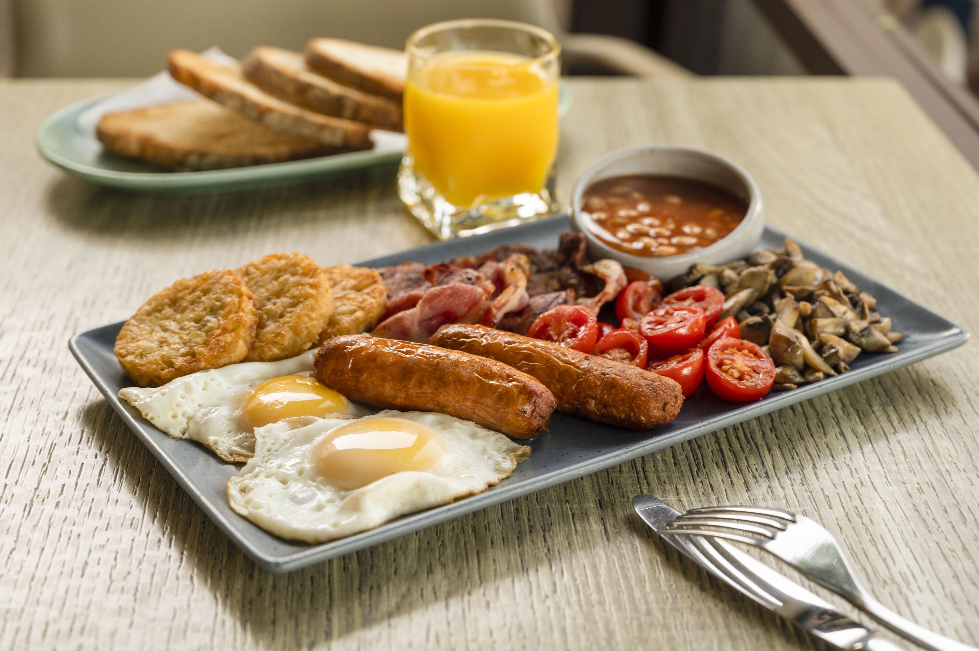 A breakfast plate with eggs, sausages, bacon, tomatoes, mushrooms, toast, and a glass of orange juice.