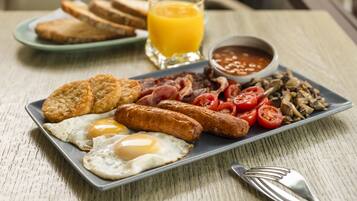 A breakfast plate with eggs, sausages, bacon, tomatoes, mushrooms, toast, and a glass of orange juice.