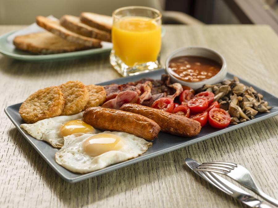 Une assiette de petit-déjeuner comprenant des œufs, des saucisses, du bacon, des tomates, des champignons, des toasts et un verre de jus d’orange.