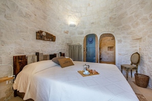 A bedroom with a stone wall, a bed with a white bedspread, a wooden headboard, a pillow with the word "Almere," a tray with a cup and croissant, and a chair with a mirror.