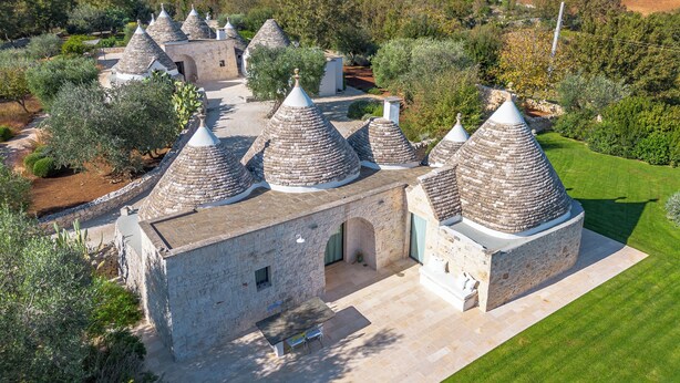 Un bâtiment traditionnel en pierre, doté de toits coniques, entouré d’oliviers et d’une pelouse bien entretenue.