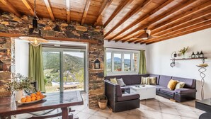 A cozy living room with a stone wall, wooden ceiling beams, a sofa set, a dining table with oranges, and a view of the outdoors. - Charming 1-bed House in Stafylos (Skopelos)