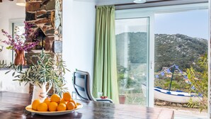 A room with a stone fireplace, a table with oranges and flowers, a chair, and a view of a boat and hills through a glass door. - Charming 1-bed House in Stafylos (Skopelos)