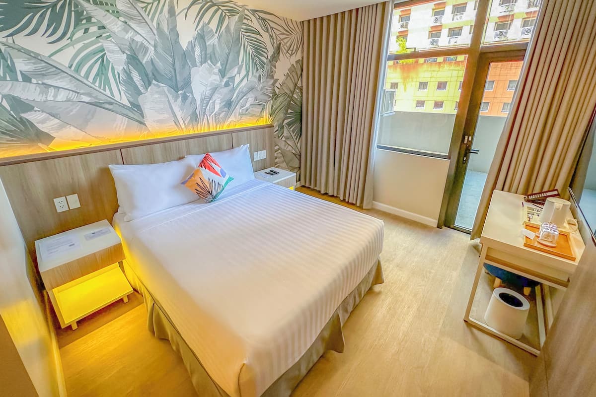 A hotel room with a bed, nightstands, a small table with a lamp, a bathroom with a toilet, and a view of buildings through the window.