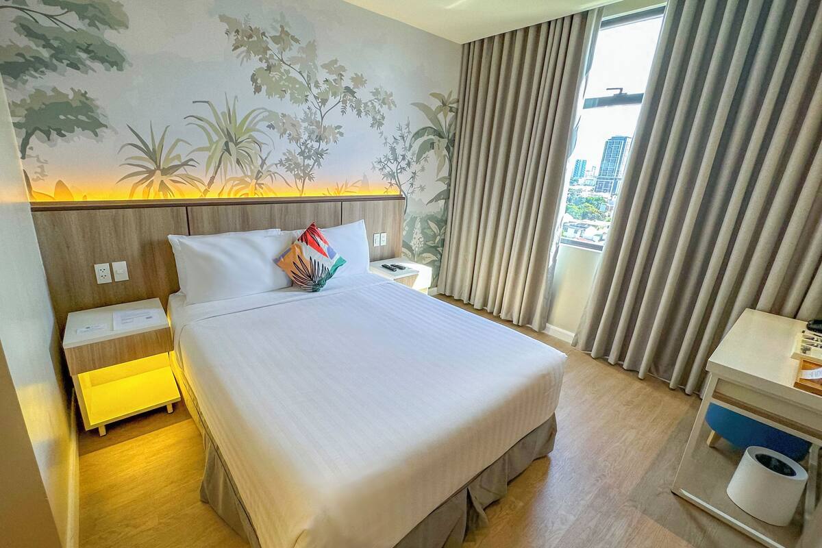 A hotel room with a bed, a nightstand, a desk, a chair, a trash bin, and a window with curtains.