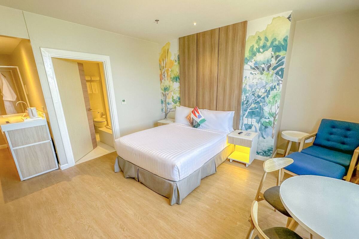 A hotel room with a bed, bedside table, chair, and a small desk with a lamp.