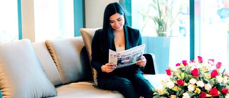 A woman sitting on a sofa reading a magazine in a well-lit room with large windows and a potted plant on a small table.