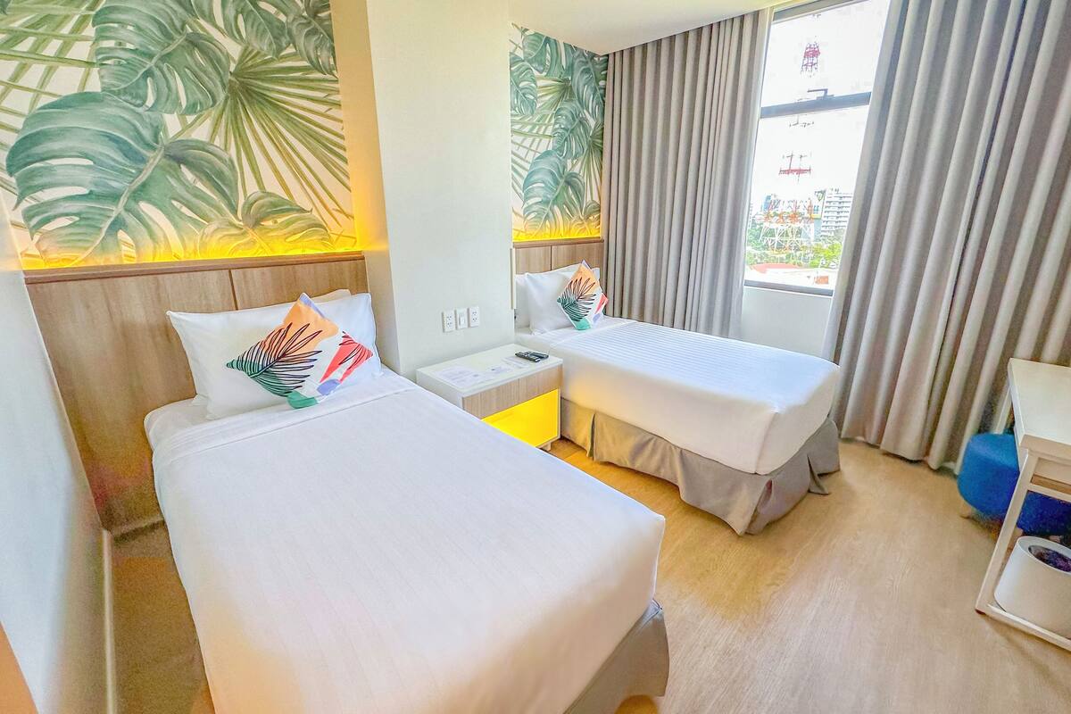 A hotel room with two beds, a nightstand, a desk, a chair, and a window with curtains.
