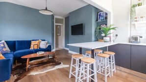 A modern living room with a blue sofa, wooden coffee table, and white bar stools. A flat-screen TV is mounted on the wall, and there is a kitchen area with a white countertop and cabinets. - Luxuriously Designed 3 Bedroom Apartment in Clapham (London)