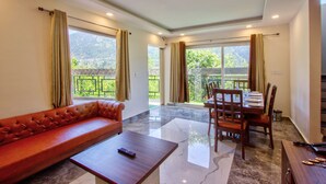 Amplia sala de estar con un sofá de cuero, una mesa de centro de madera y una mesa de comedor preparada para cuatro personas. Grandes ventanales ofrecen vistas a zonas verdes y un balcón.