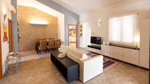 A modern living room with a sofa, dining table, and chairs, a kitchen area with cabinets and a TV, and a brick wall.