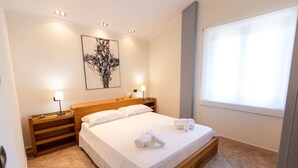 A neatly made bed with white linens and towels, a wooden headboard, a bedside table with a lamp, and a large window with blinds.