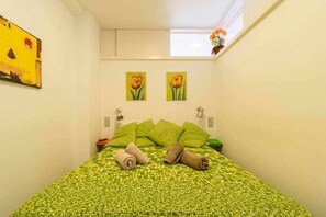 Ein kleines, gemütliches Schlafzimmer mit einer zarten, blumigen Tagesdecke, zwei grünen Kissen und zwei zusammengerollten Handtüchern auf dem Bett. An der Wand sind zwei Lampen angebracht und darüber hängen zwei Blumenbilder.