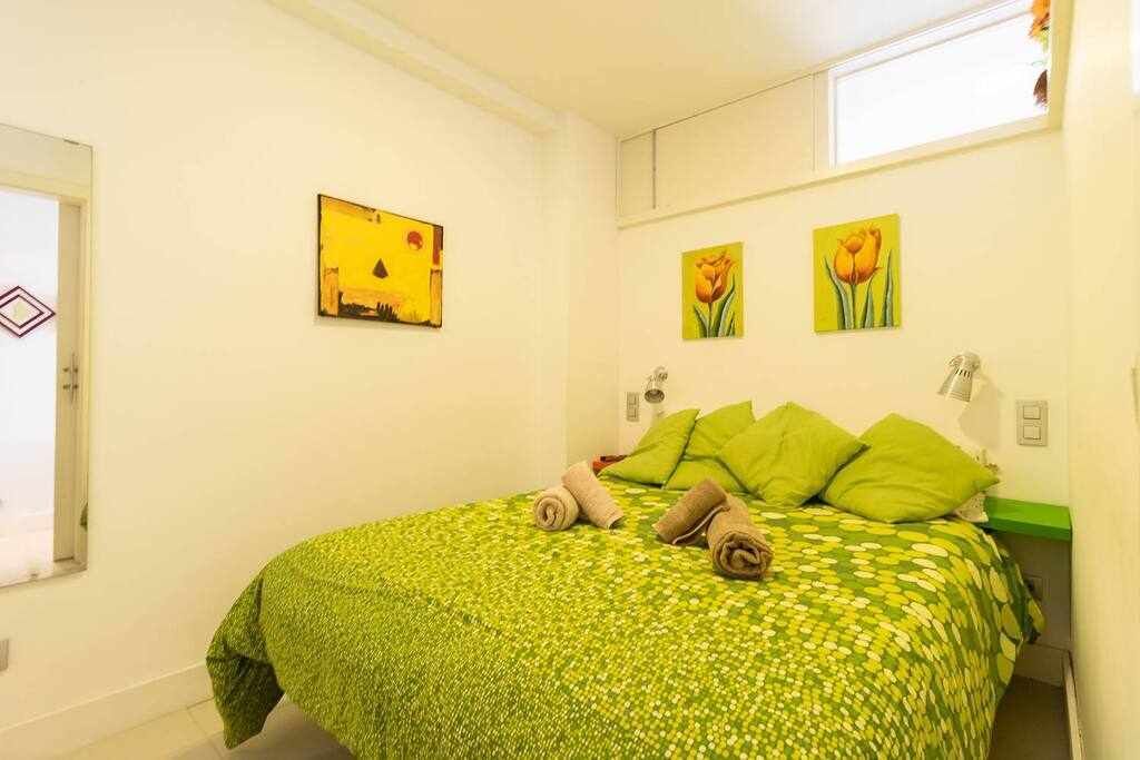 A room with a bed, green floral bedspread, two green pillows, and two rolled towels. There are two wall-mounted lamps and two paintings of tulips.