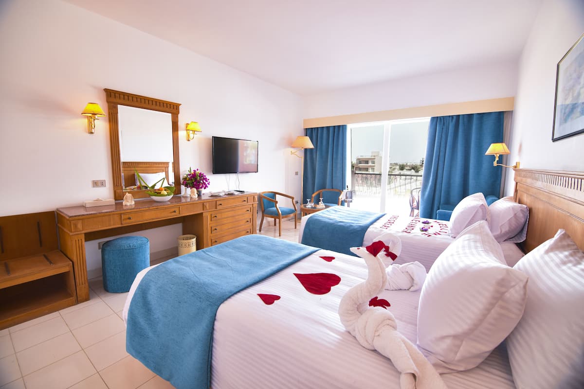A hotel room with two beds, a desk, a chair, a television, and a balcony with blue curtains.