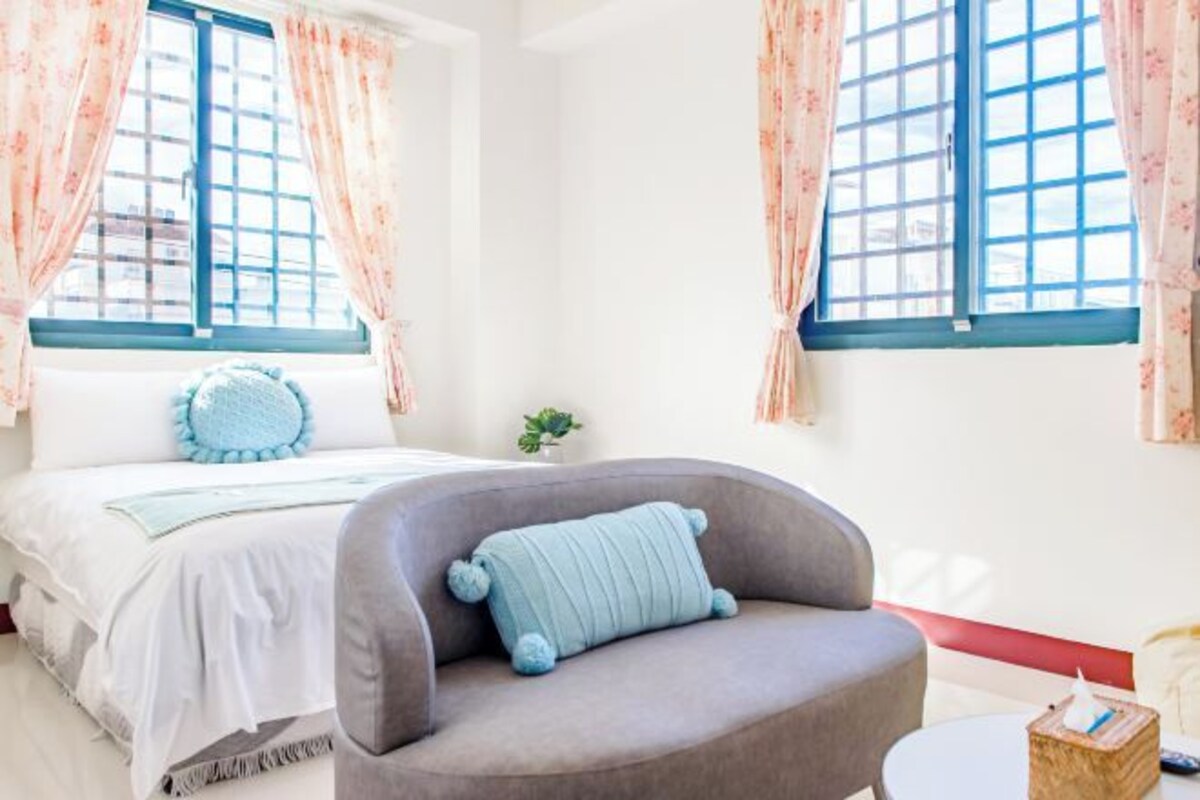 A bedroom with a bed, a grey armchair, and two windows with floral curtains.