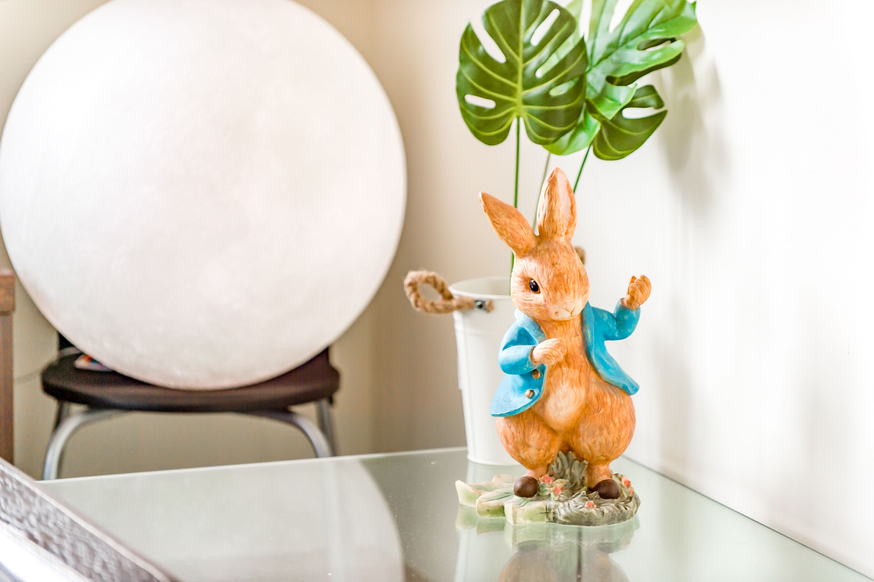 A figurine of a rabbit on a glass table with a white cup and a large green plant in the background.