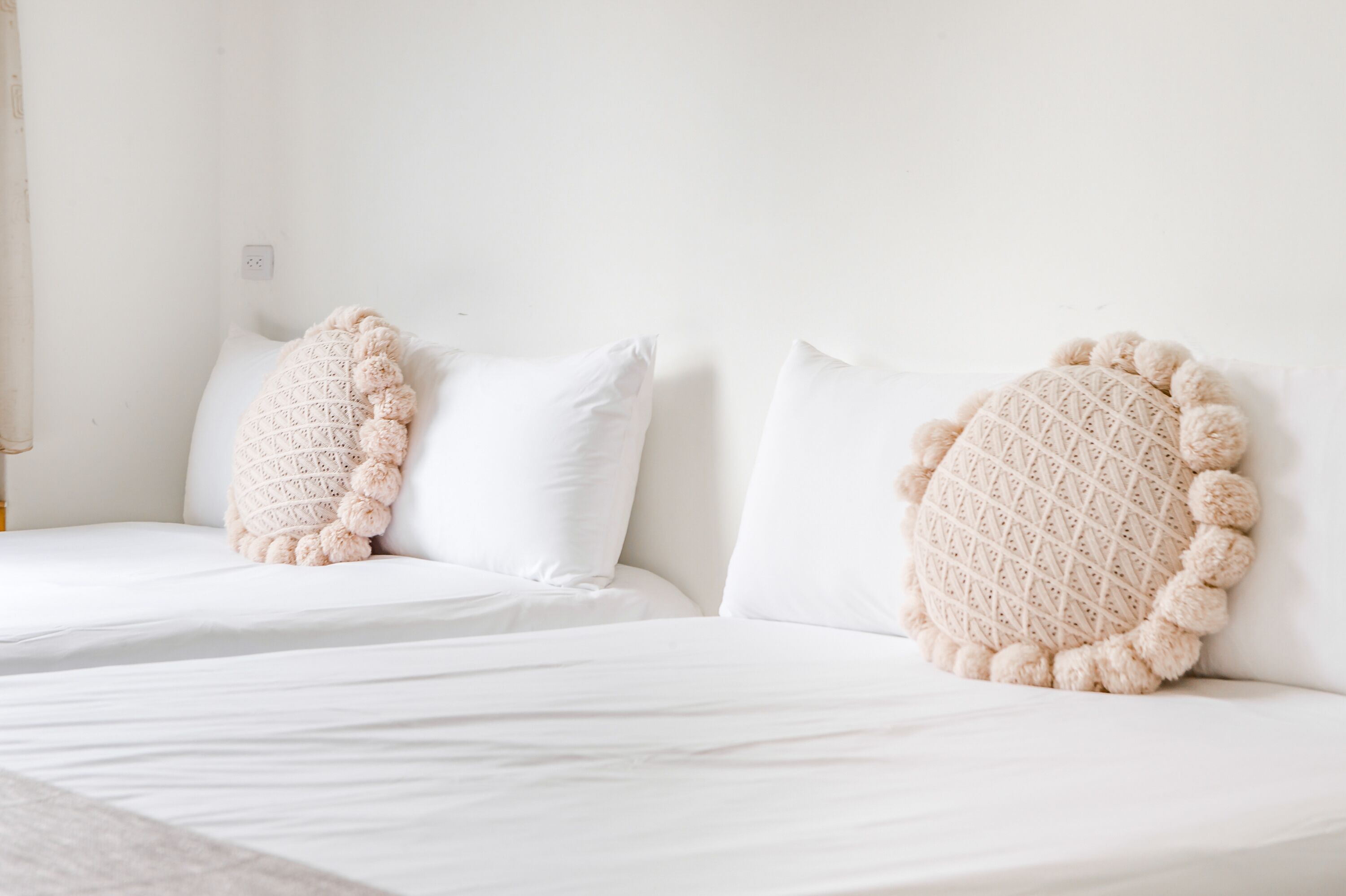 A hotel room with two beds, each with a white mattress and a textured beige throw pillow.