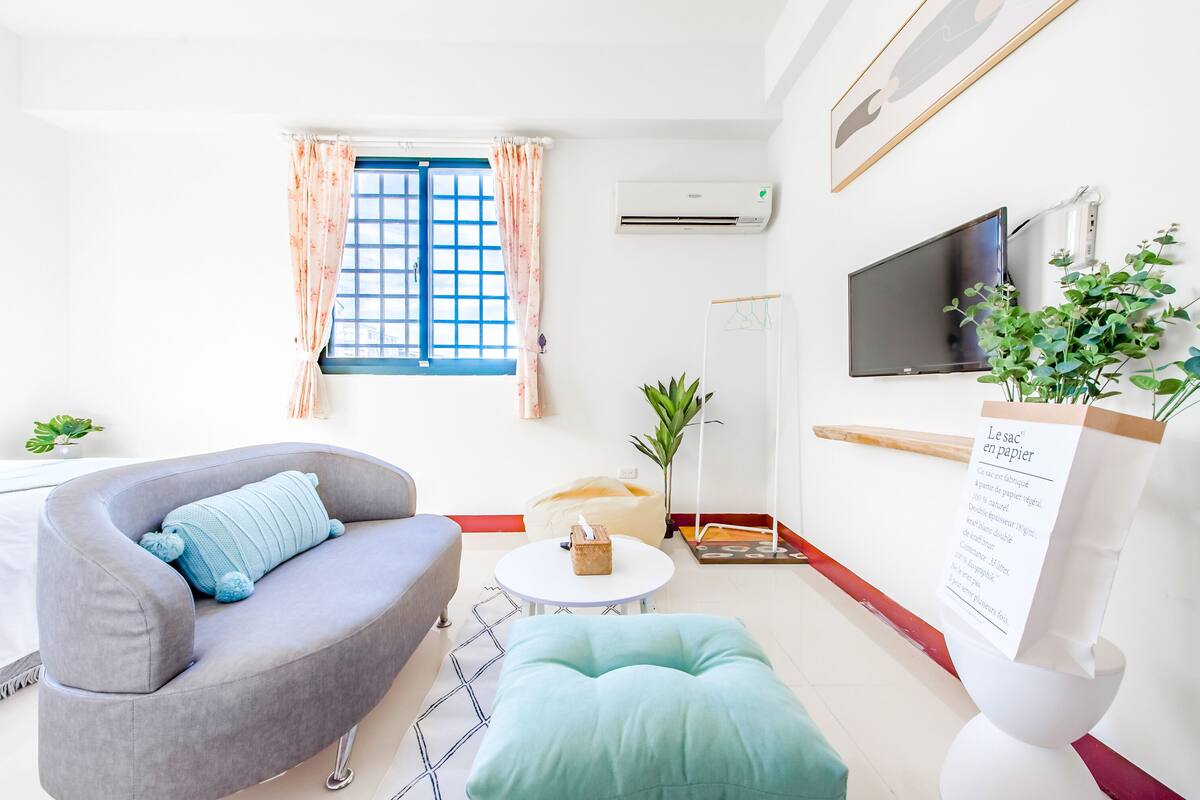 Un saló modern amb un sofà gris, una taula rodona petita, un seient de ploma, un gran finestral amb cortines, un televisor de pantalla plana i una pissarra blanca.