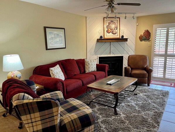 A living room with a red sofa, a plaid armchair, a coffee table, a fireplace, and a ceiling fan. - Golf Course Front Condo | Ruidoso Getaway w/ (Ruidoso)
